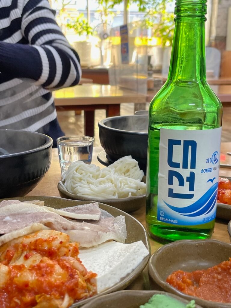Daesun green soju bottle with boiled pork, noodles and kimchi on a sunlit Korean restaurant table